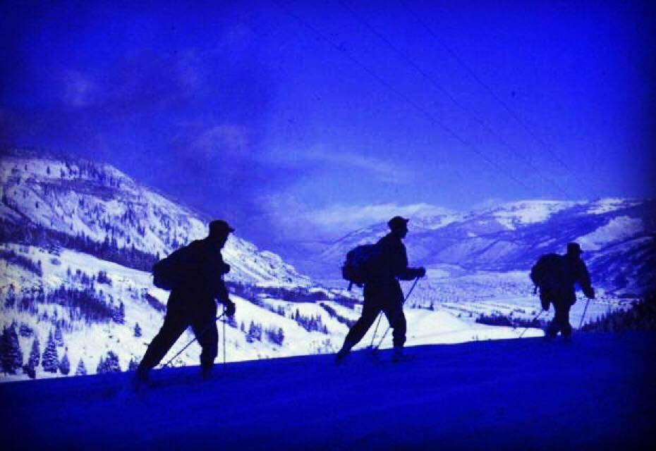 WWII-era 10th Mountain Division alpine troops silhouetted on a snowy ridge, carrying full packs and skis.